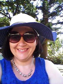 Smiling light skinned women, wearing dark sunglasses, a white wide brimmed hat with trees in the background